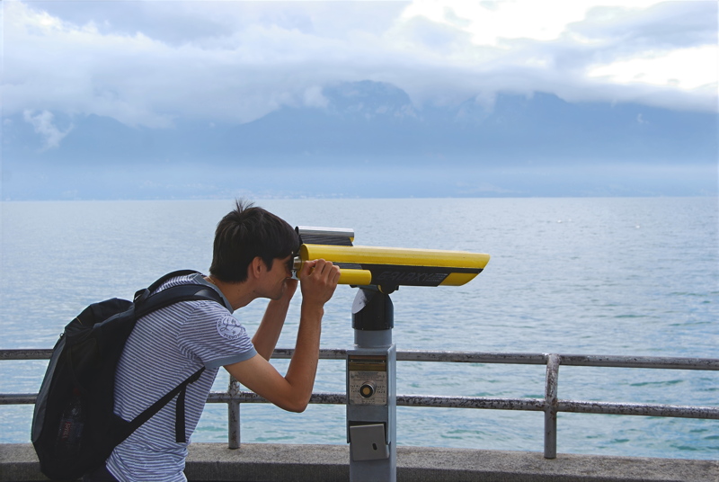 A person looking through a yellow public coin-operated observation telescope by a large body of water with distant mountains under a cloudy sky